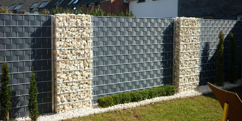 Doppelstabmatten-Sichtschutzzaun aus weiß pulverbeschichteten Matten und Pfosten, mit blauen Sichtschutzstreifen und Steineinlage